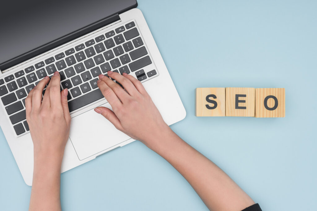 Hands typing on laptop keyboard with wooden blocks spelling SEO on light blue background