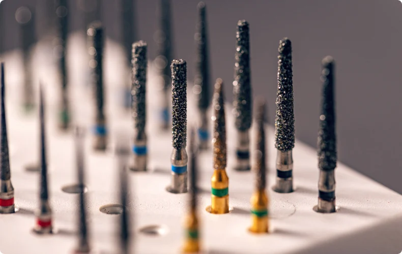 The image shows a set of diamond dental burs, which are rotary instruments used primarily in dentistry for cutting, grinding, and polishing hard tooth structures or dental materials. The burs vary in shape and size and are marked with colored bands indicating grit or intended use. These tools are typically mounted in a holder tray, as seen in the image.